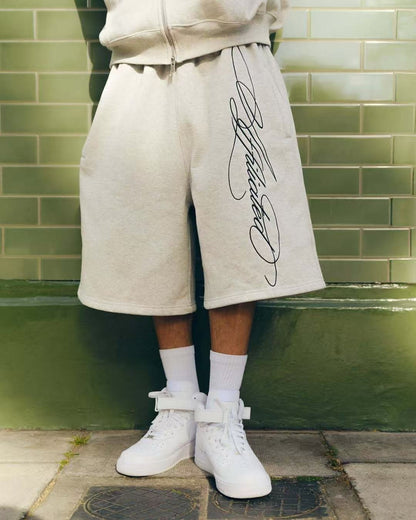 Person wearing beige shorts with black script print, white socks, and white high-top sneakers against green tiled wall