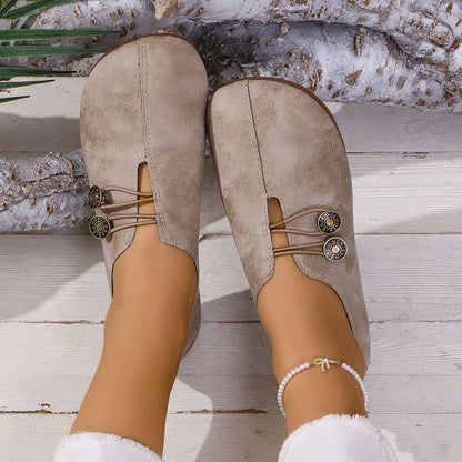 Beige suede slip-on shoes with button details worn on feet, styled with pearl anklet on wooden floor