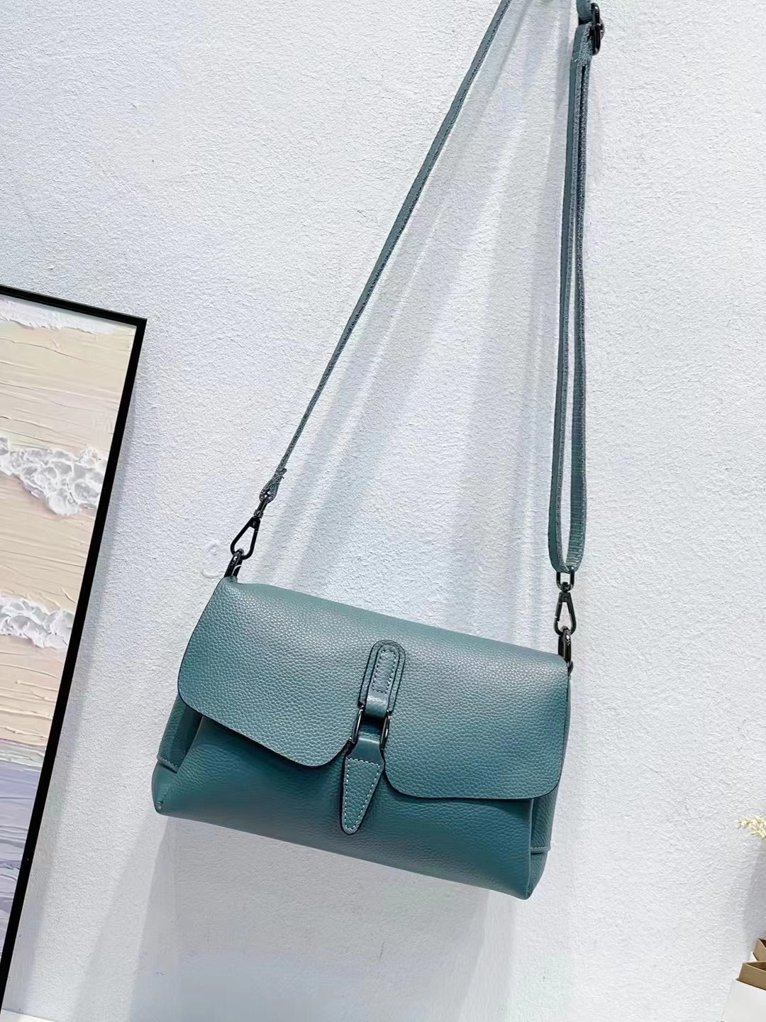 Blue leather crossbody bag with flap and buckle hanging on white textured wall