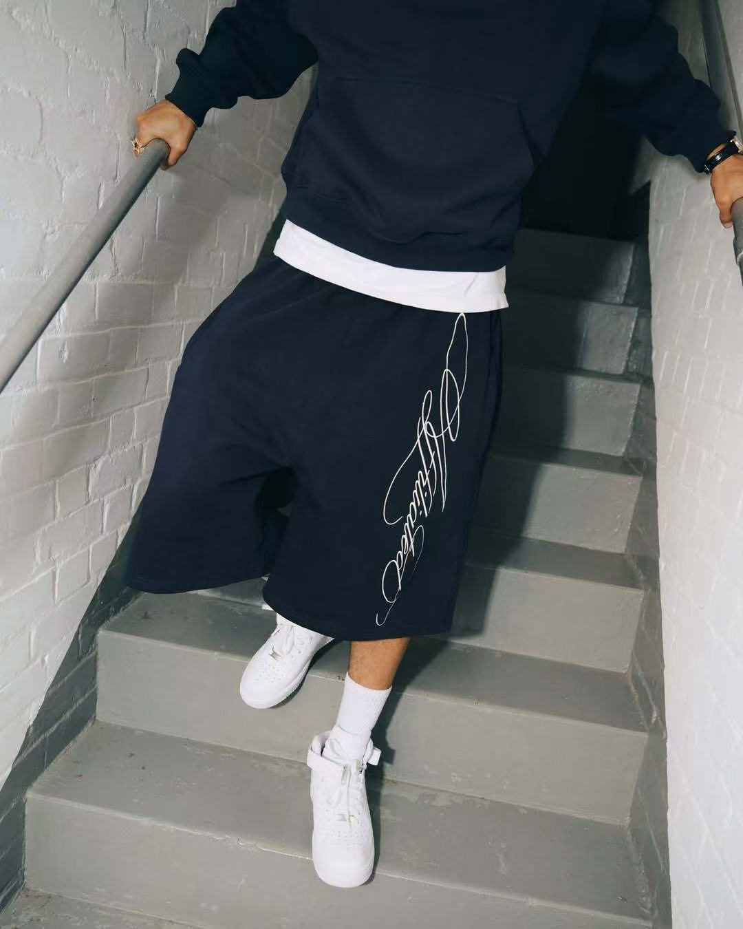 Man in black oversized shorts with white script, black hoodie, white sneakers, descending gray stairs