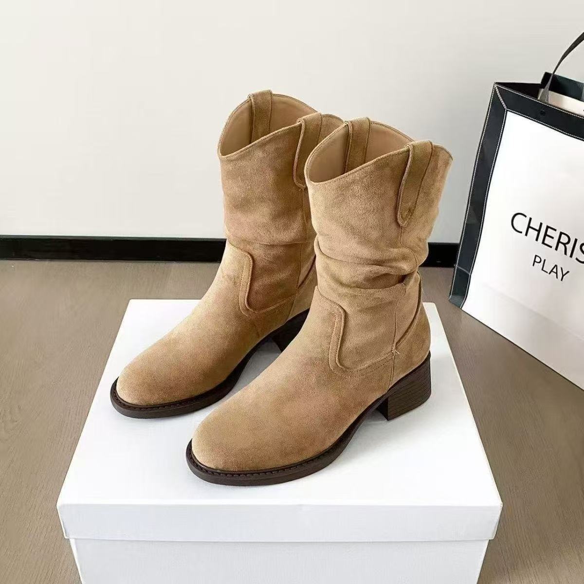 Pair of tan suede mid-calf boots with block heels on a white box, next to a Cherish Play bag