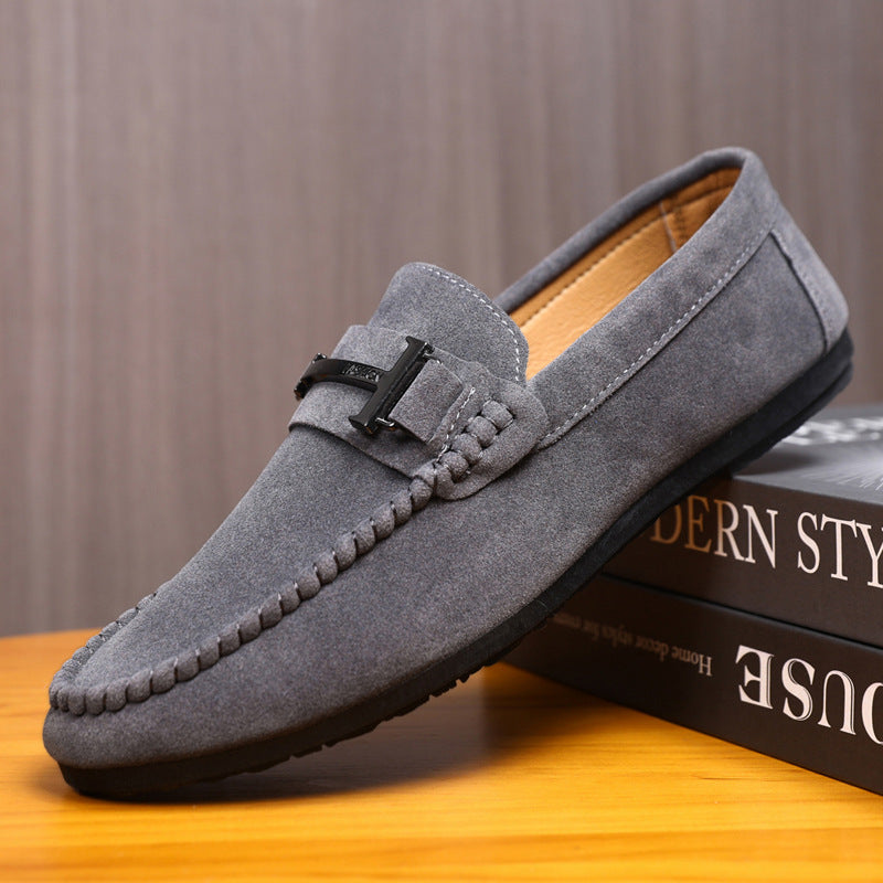 Gray suede men's loafer with black metal buckle stitching detail on wooden surface and books
