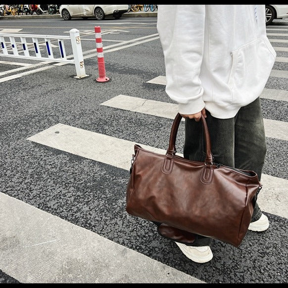 Person in white hoodie and dark pants holding a large brown leather duffle bag on a city crosswalk