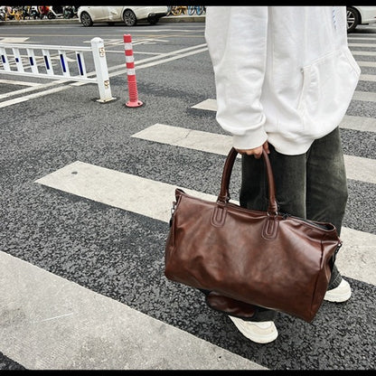 Person in white hoodie and dark pants holding a large brown leather duffle bag on a city crosswalk