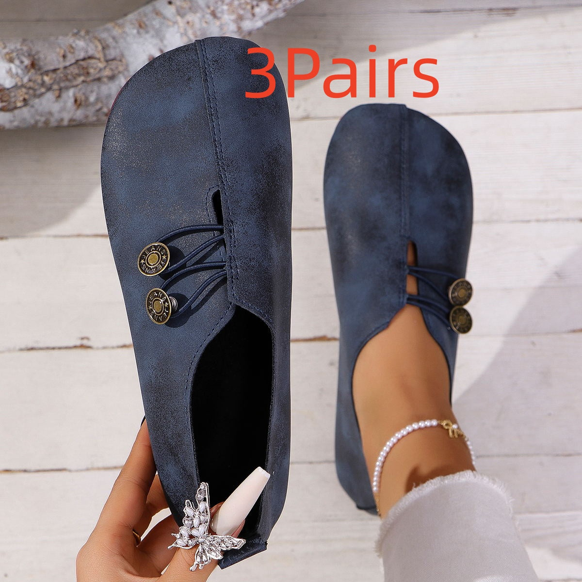 Pair of navy blue minimalist slip-on flat shoes with decorative buttons, worn on wooden floor