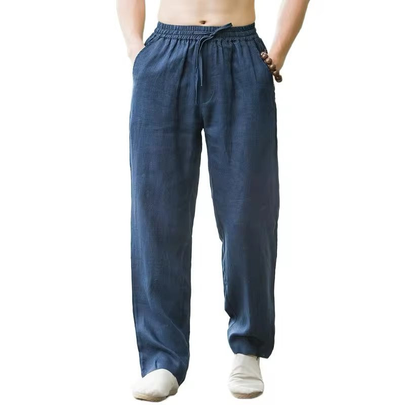 Man wearing blue loose linen pants with elastic waist and drawstring, hands in pockets on white background