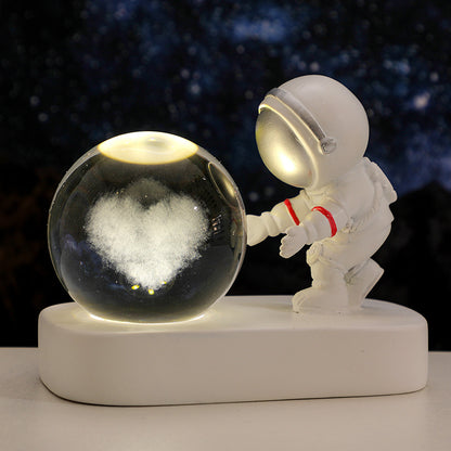 White astronaut figurine reaching towards illuminated glass crystal ball with heart-shaped snow inside