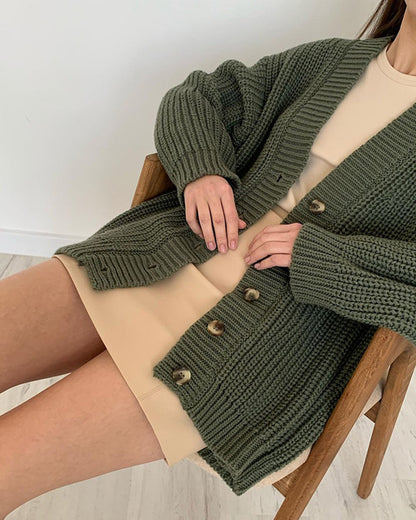 Woman wearing an olive green chunky knit cardigan with buttons over a beige dress while sitting on a wooden chair