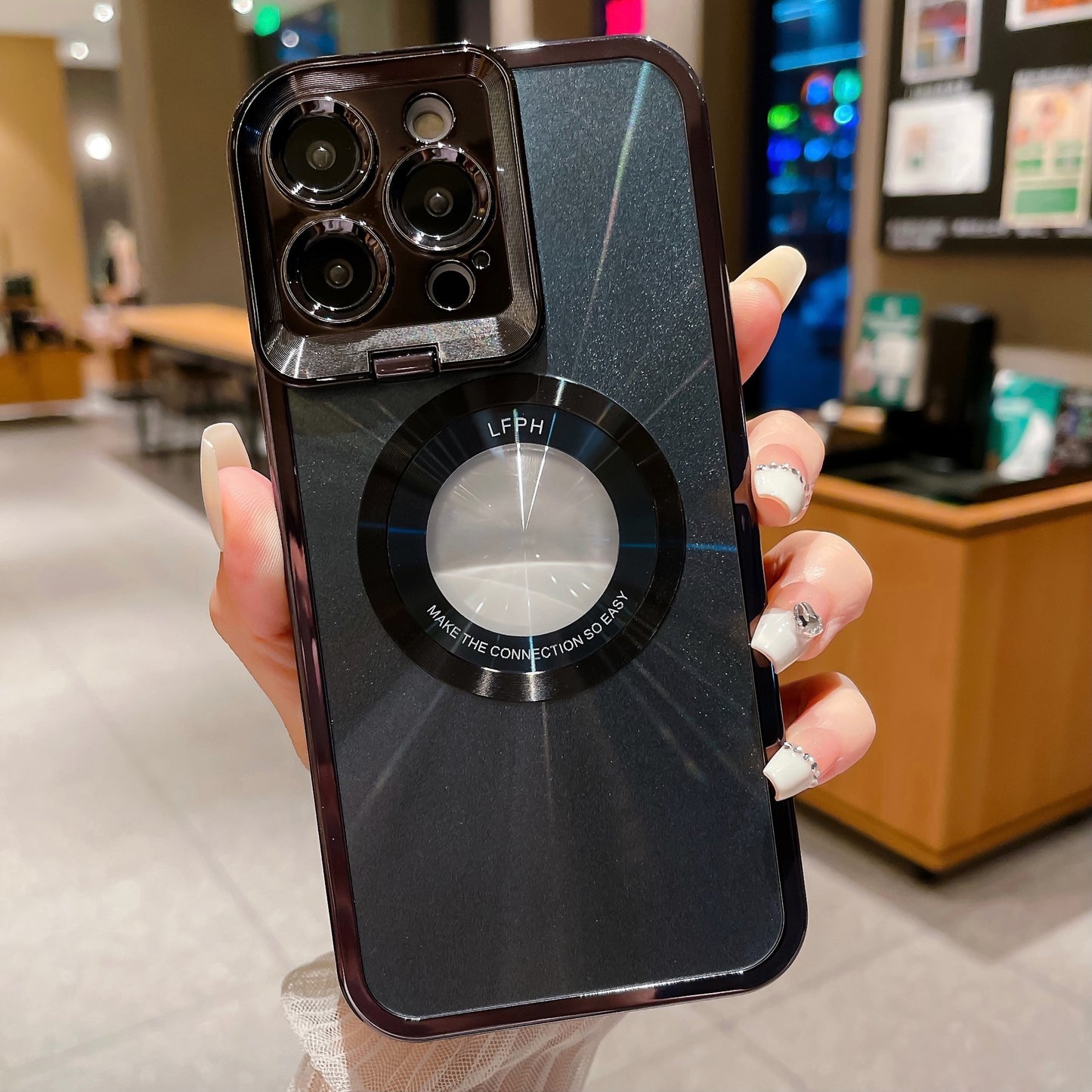 Hand holding black glossy phone case with triple camera and circular design in indoor cafe setting