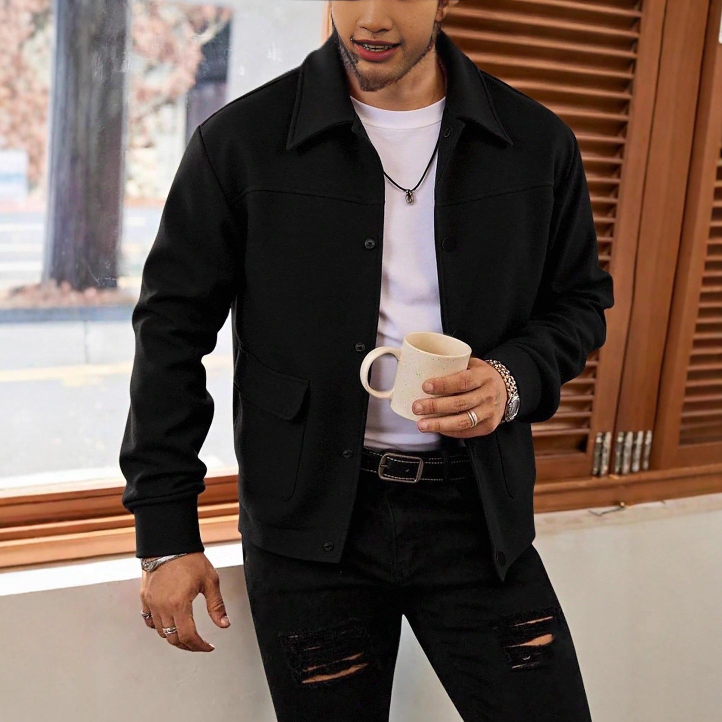 Man wearing black jacket, white t-shirt, and ripped black jeans holding a beige mug indoors