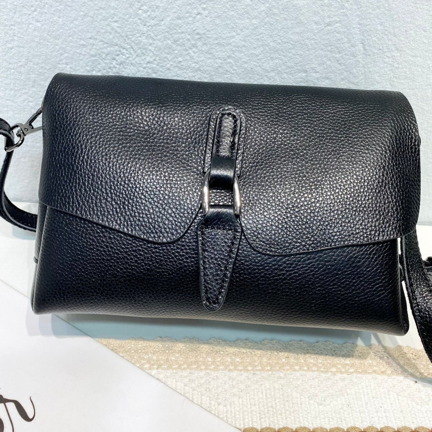 Black textured leather crossbody bag with silver buckle and strap on white surface