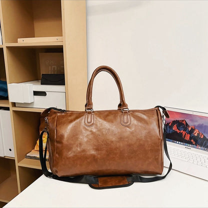 Brown leather duffel bag with handles and adjustable strap on white table near wooden shelves and laptop