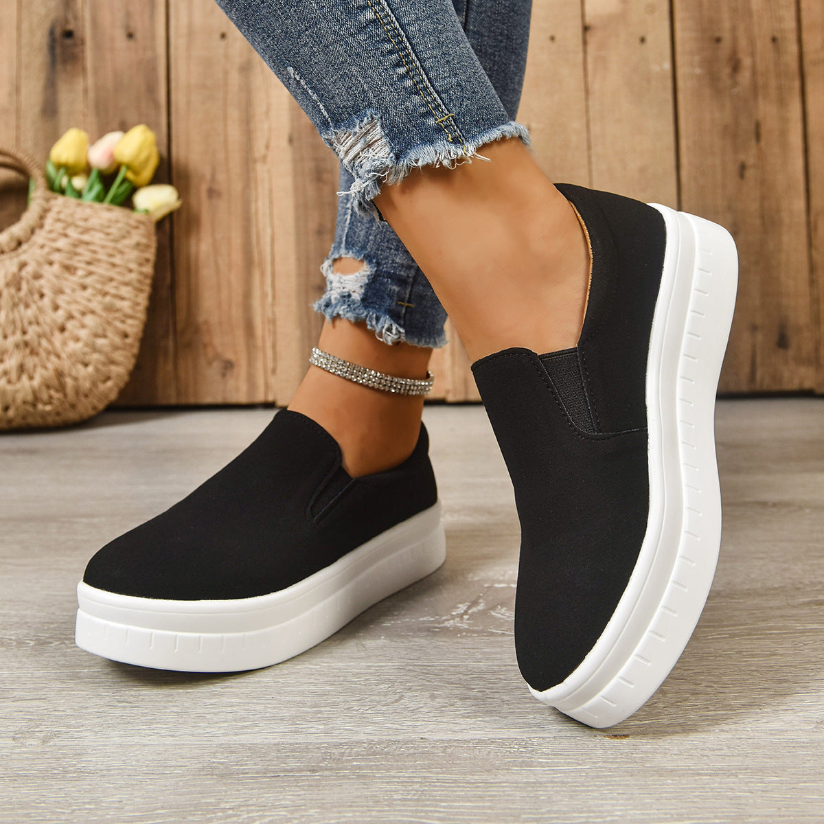 Black slip-on platform sneakers with white soles worn with ripped blue jeans and anklet on wood floor