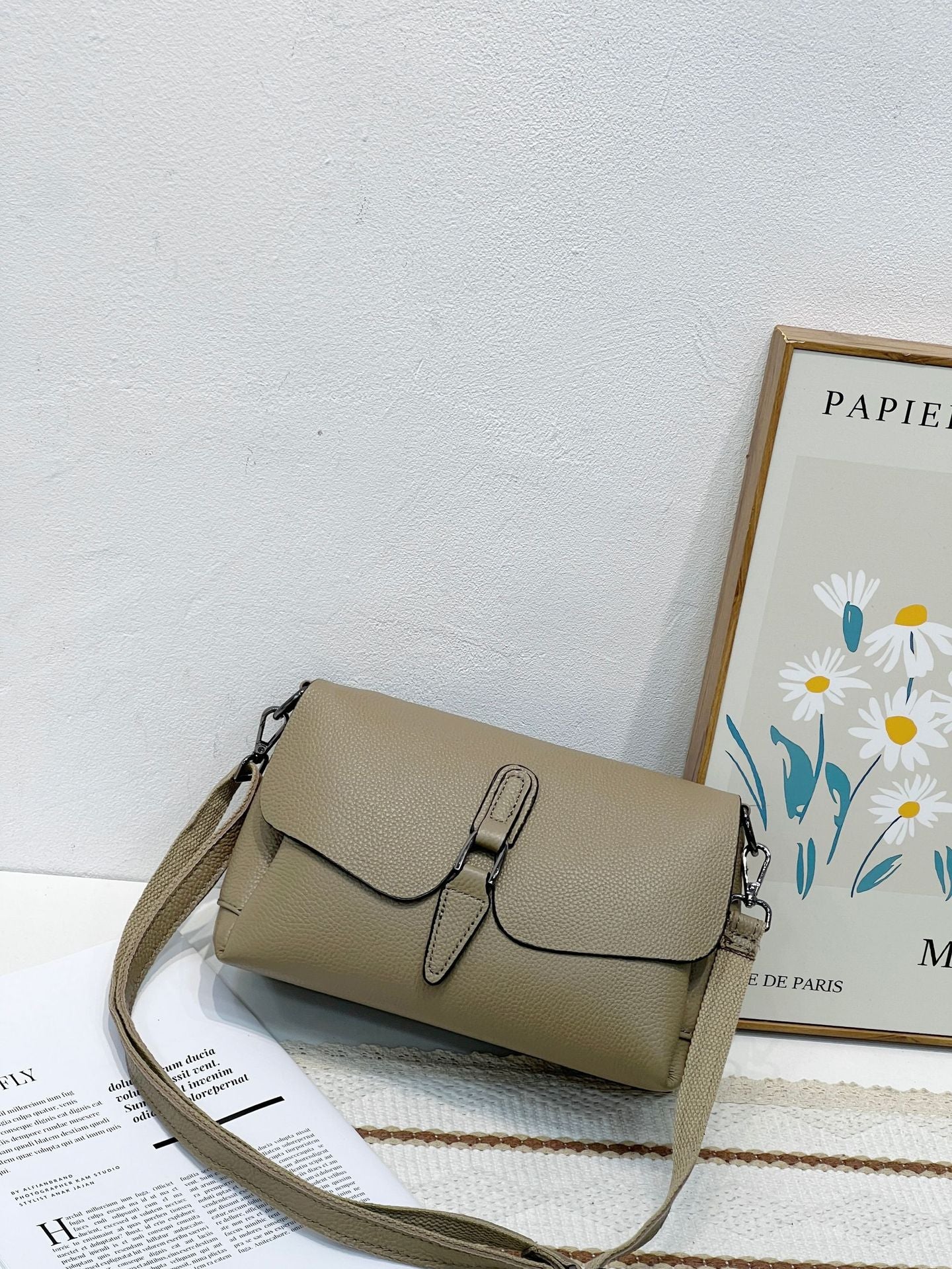 Beige textured leather shoulder bag with buckle detail on white surface, next to floral framed artwork