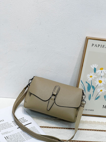 Beige textured leather shoulder bag with buckle detail on white surface, next to floral framed artwork