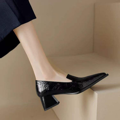 Close-up of a foot wearing a black pointed-toe block heel shoe with a textured finish, beige background