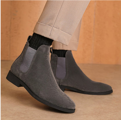 Men's gray suede Chelsea boots paired with black socks and beige pants against a minimalist background