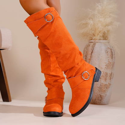 Bright orange suede knee-high boots with silver buckle accents worn indoors by a model