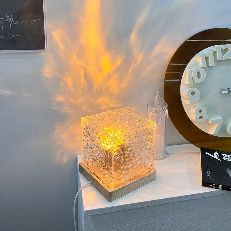 Modern glass cube table lamp glowing with warm light beside a large wall clock on a white shelf