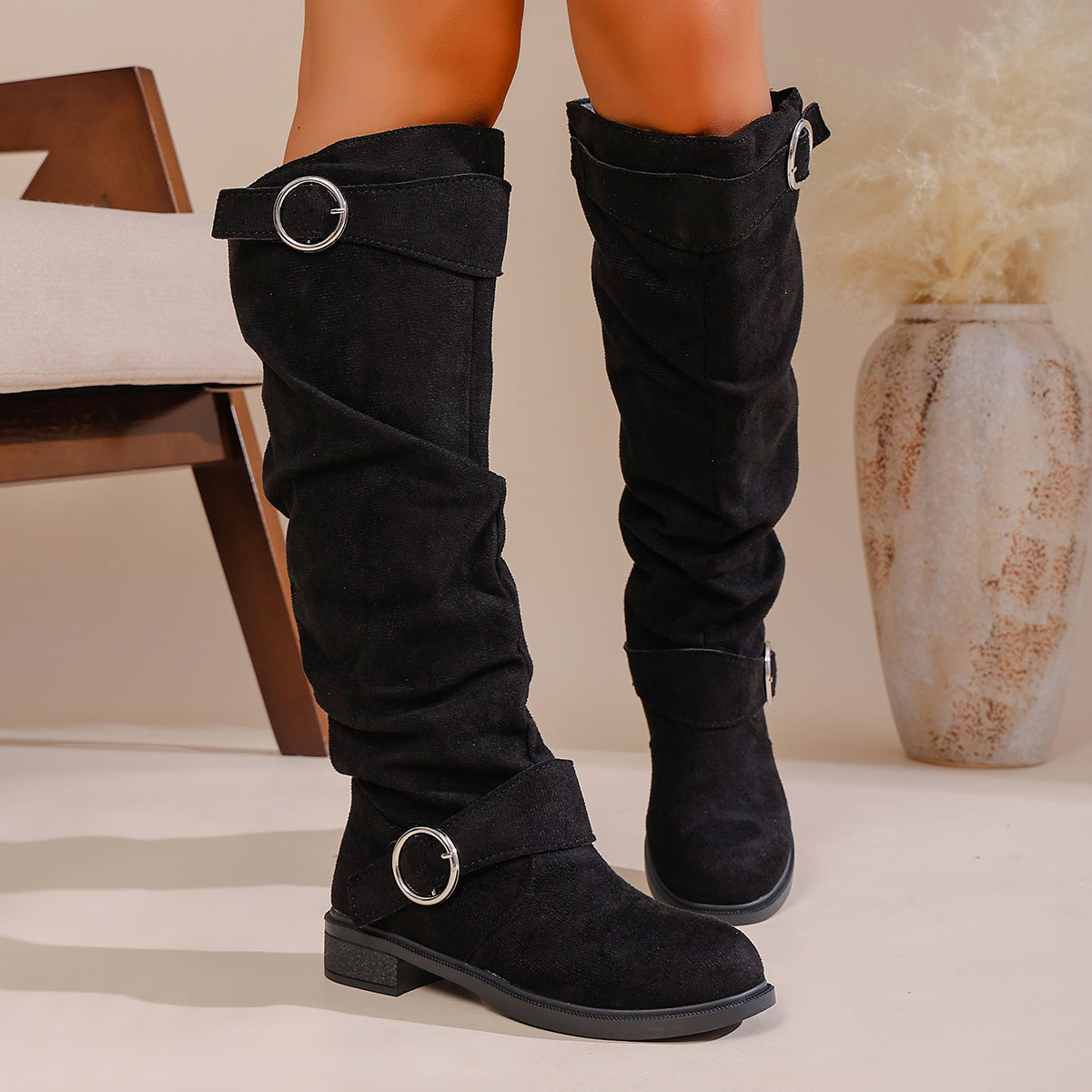Black suede knee-high boots with silver buckle straps worn indoors near chair and decorative vase