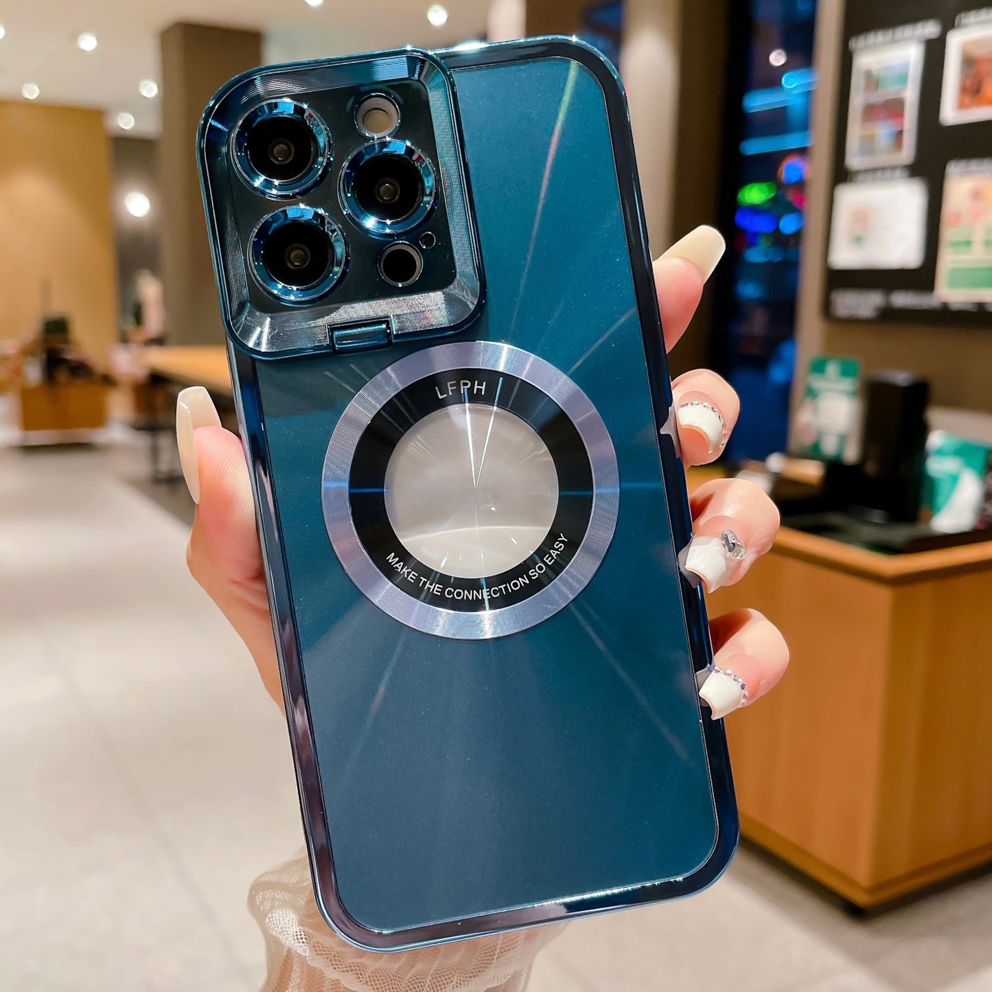 Hand holding blue smartphone with camera lenses and circular metal ring case in cozy indoor cafe