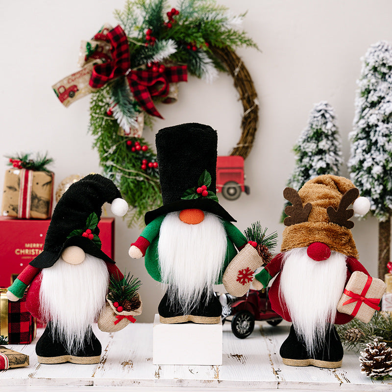 Three Christmas gnome decorations with long white beards and festive hats on wooden surface, pine wreath and snowy trees background