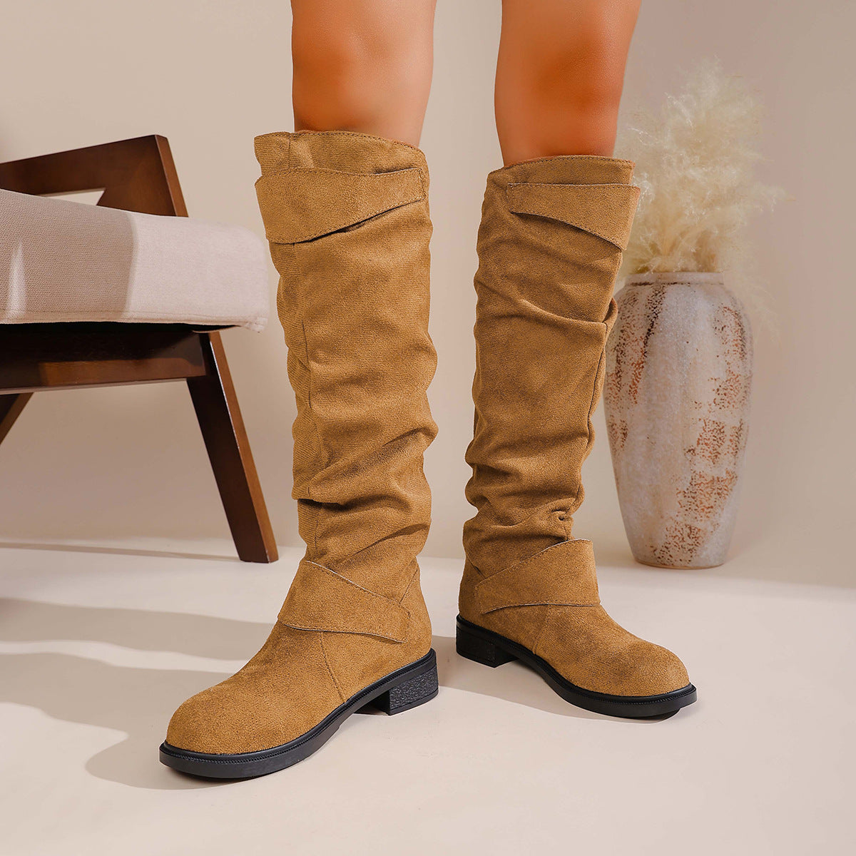 Women's tan suede knee-high boots with strap detail worn indoors near chair and vase