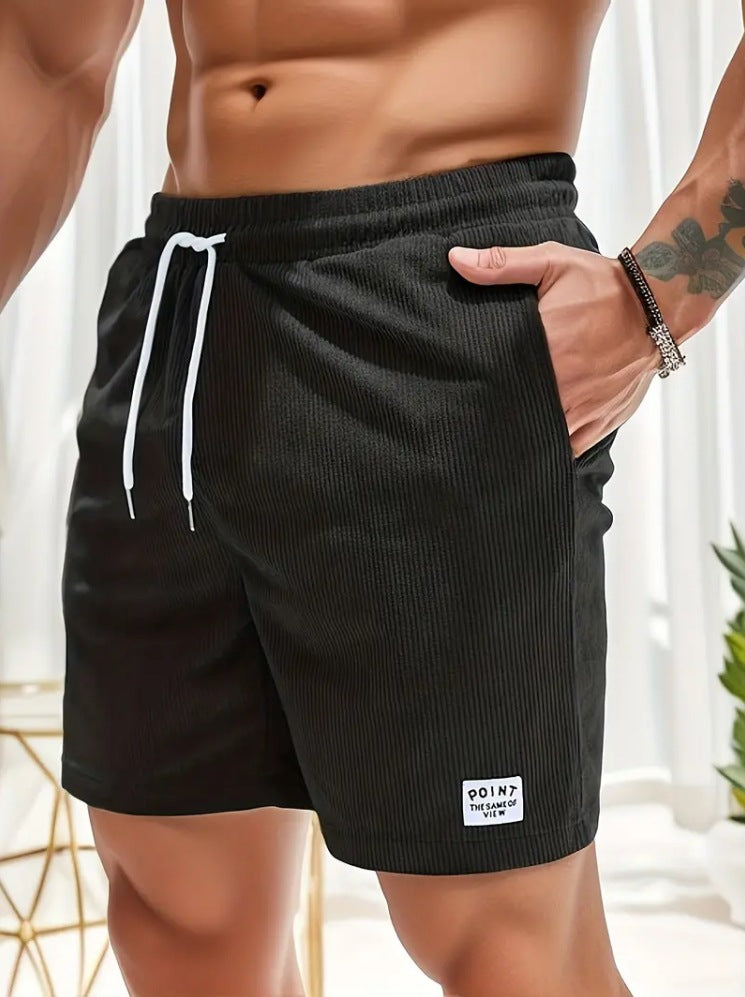 Male torso wearing black ribbed shorts with white drawstring and patch, hand in pocket, indoor bright setting