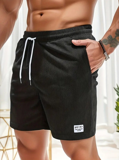 Male torso wearing black ribbed shorts with white drawstring and patch, hand in pocket, indoor bright setting