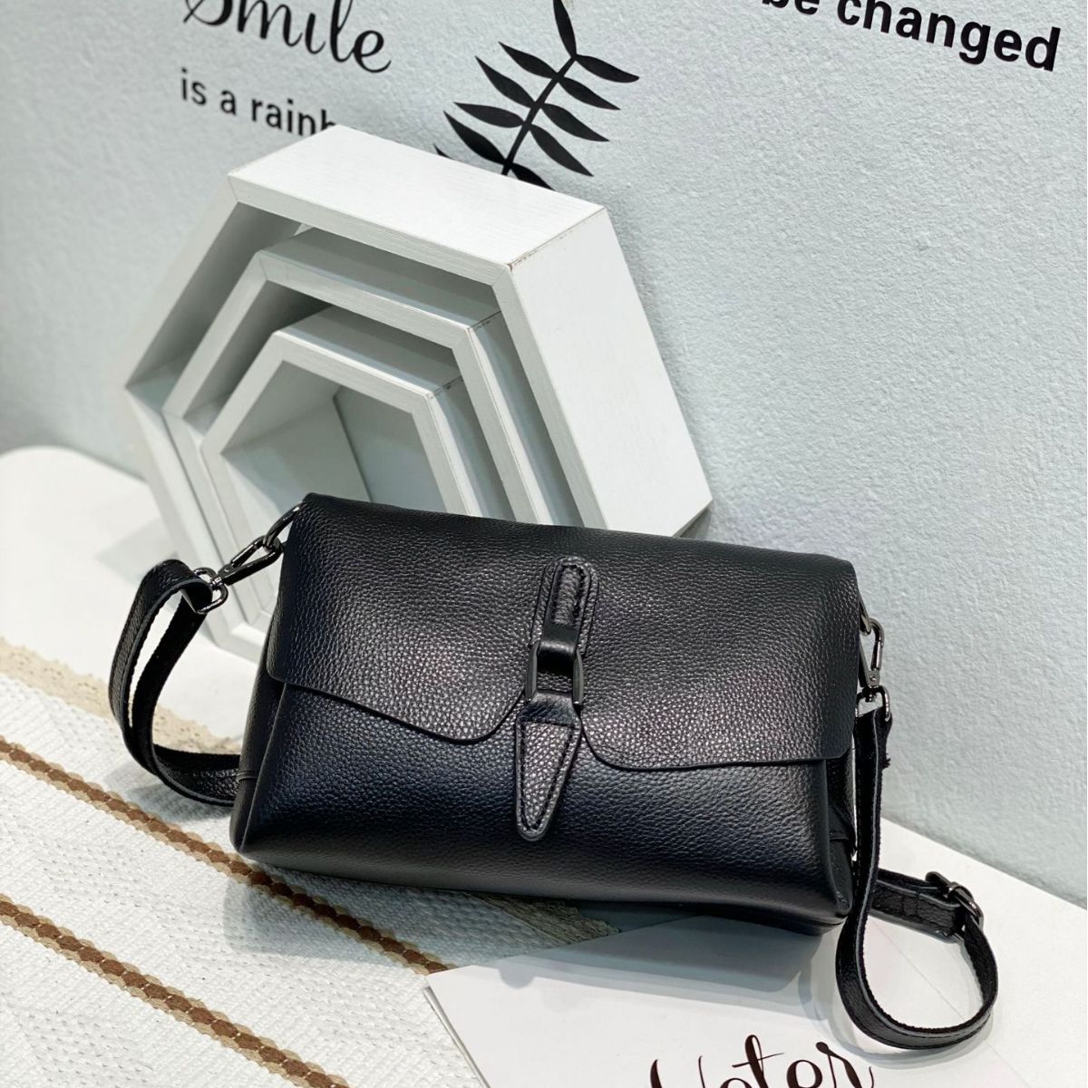 Black textured leather crossbody bag on white surface with geometric wall decor and text background