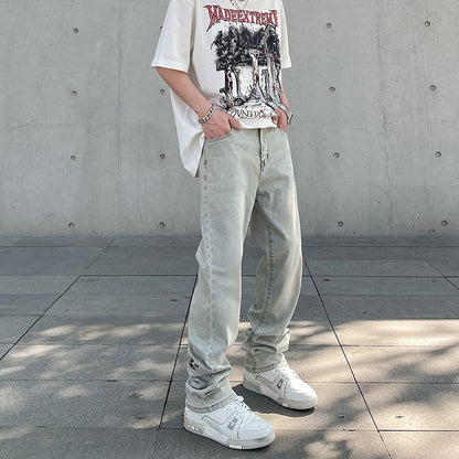 Man wearing light wash baggy jeans, white graphic t-shirt, silver accessories, and white sneakers against concrete wall