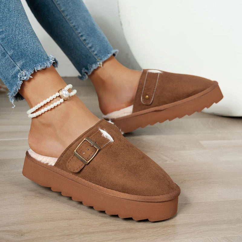 Brown suede slip-on slippers with buckle and faux fur lining on feet wearing pearl anklet and frayed jeans