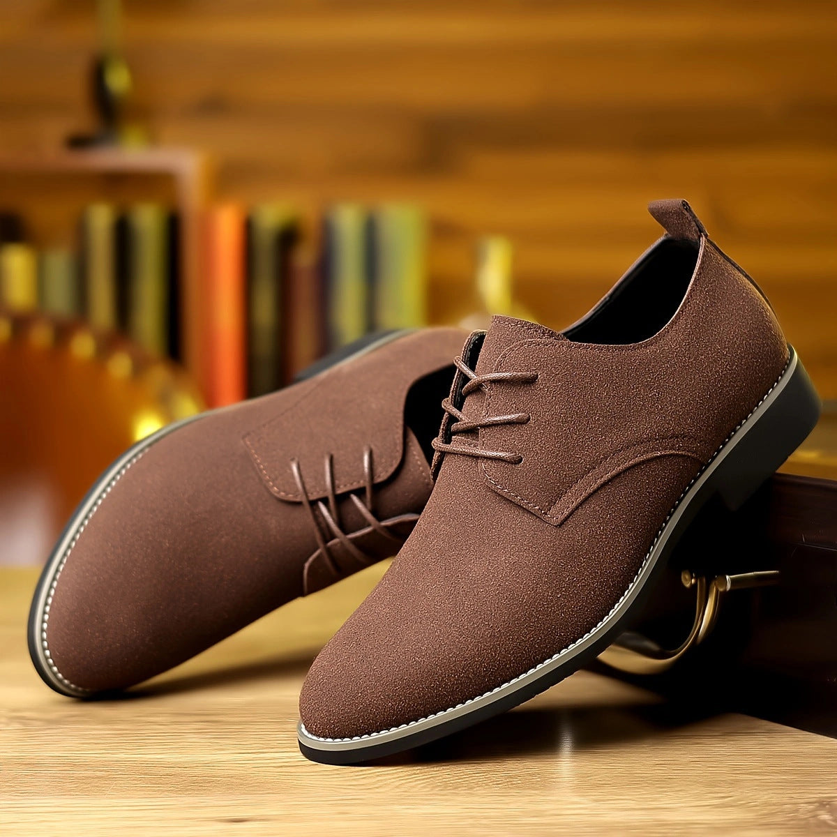 Pair of brown suede men's dress shoes with black soles on wooden surface and blurred bookshelf background