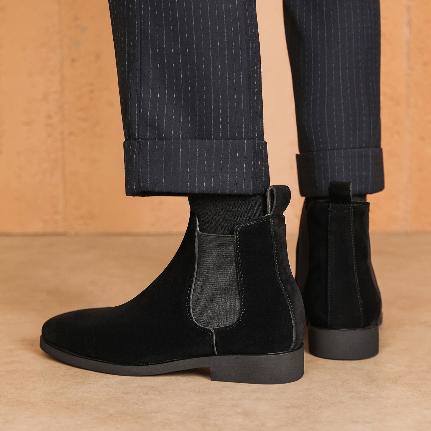 Black suede Chelsea boots worn with dark pinstripe cuffed pants on a tan floor and wall