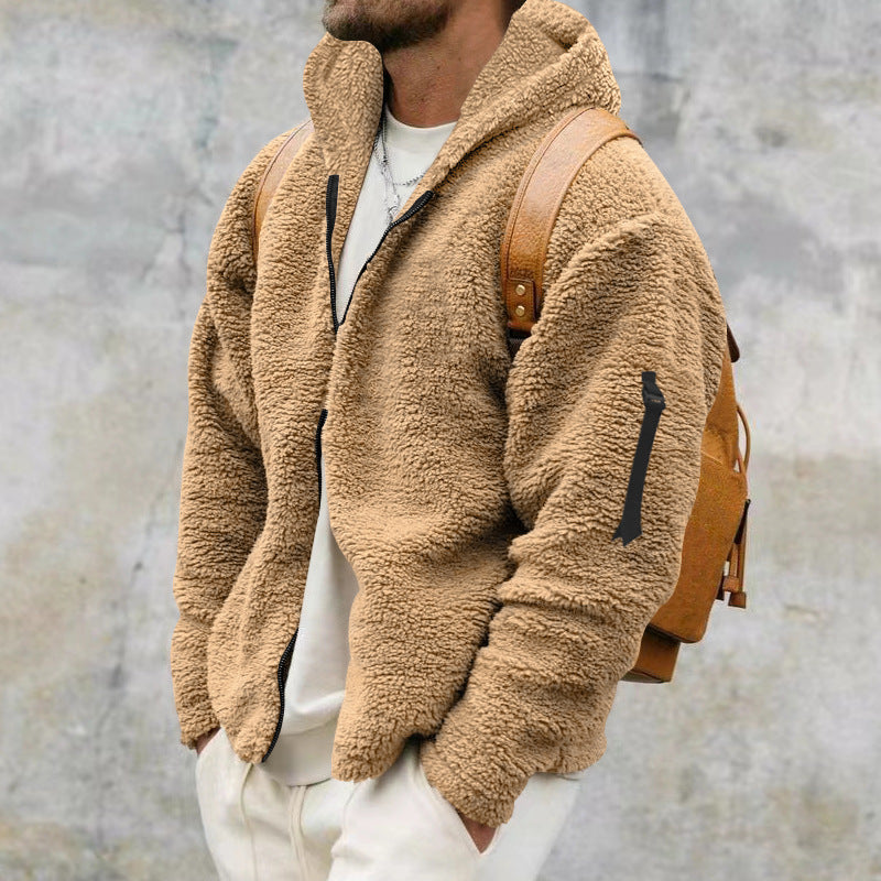Man wearing tan sherpa fleece jacket with black zipper and brown leather backpack against gray wall