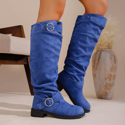 Blue suede knee-high boots with silver buckle detailing worn indoors near chair and decorative vase