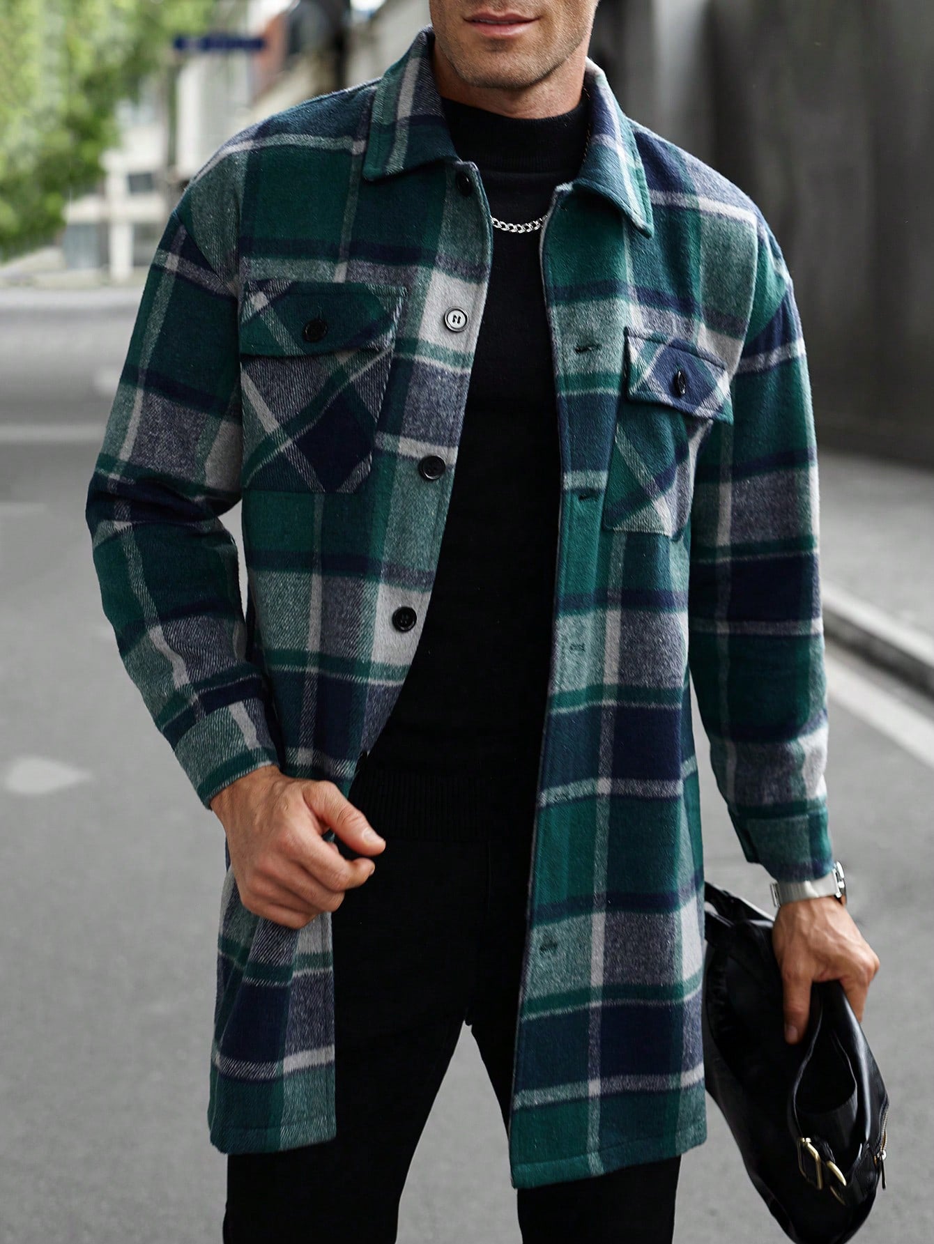 Man wearing green and blue plaid long jacket over black shirt, holding black leather bag outdoors