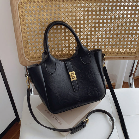 Black leather handbag with gold clasp and detachable shoulder strap on white surface with rattan chair background