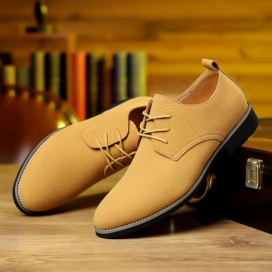 Pair of tan suede men's dress shoes with black soles on wooden surface and bookshelf background