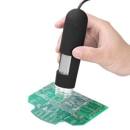 Hand holding black USB digital microscope inspecting green circuit board with LED illumination