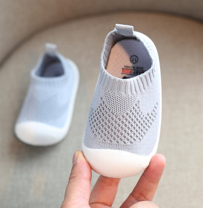 Hand holding grey toddler slip-on shoe with breathable knit upper and flexible white sole