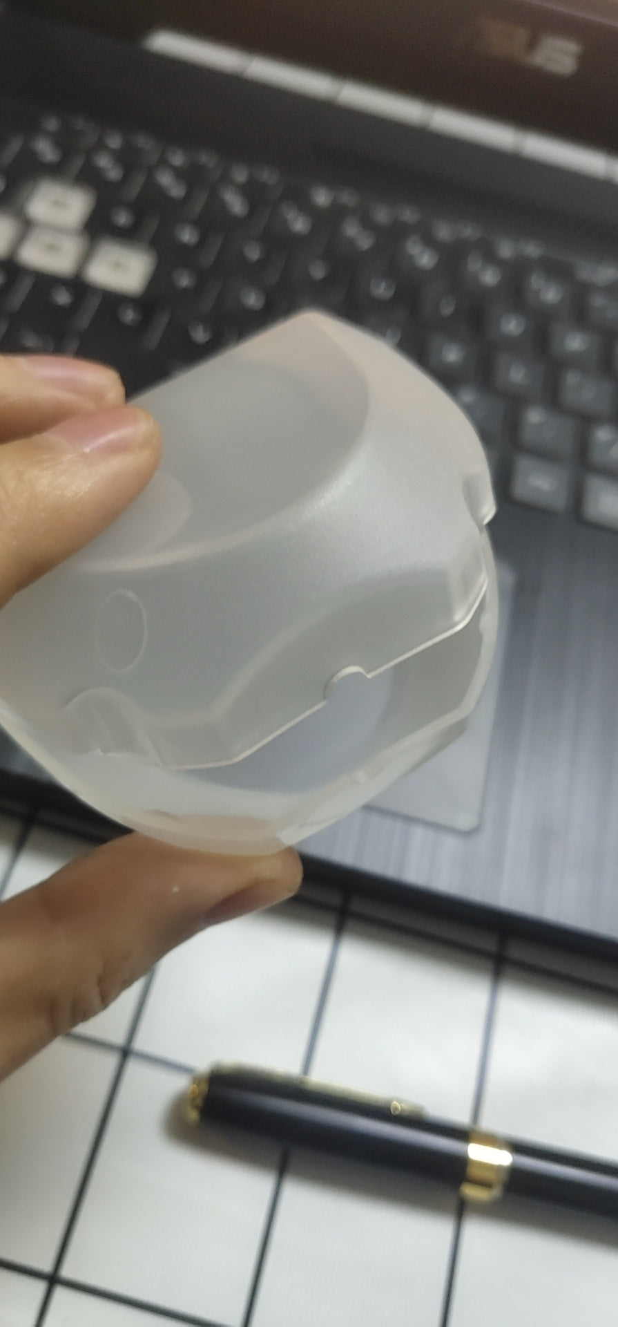 Hand holding a translucent plastic case for electric toothbrush head with a laptop keyboard and pen in background