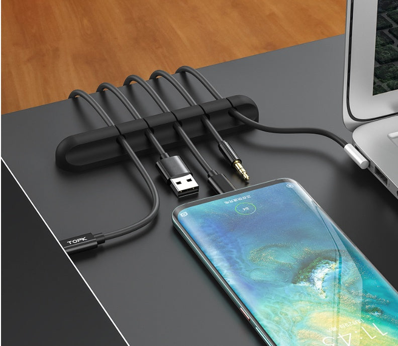 Black silicone cable organizer holding multiple cables on a desk next to a smartphone and laptop