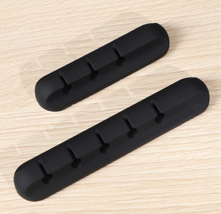 Two black silicone cable organizers with multiple slots on a light wood surface for desk wire management
