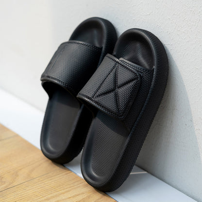 Black EVA platform slippers with wide adjustable straps leaning against white textured wall on wooden floor