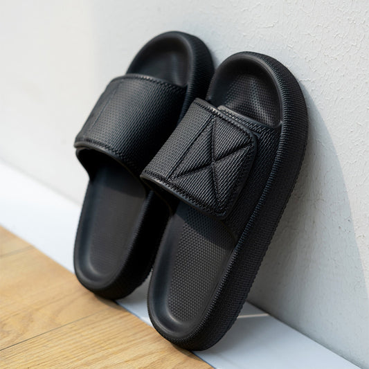 Black EVA platform slippers with wide adjustable straps leaning against white textured wall on wooden floor