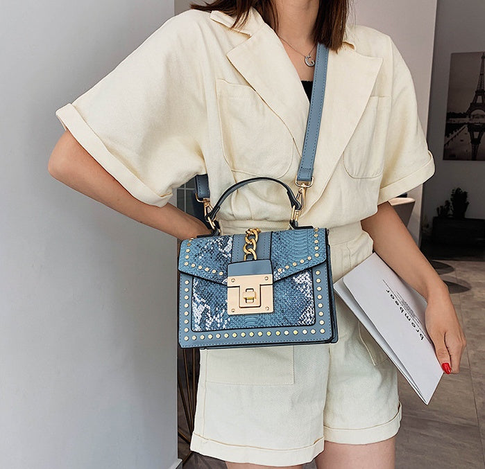 woman holding blue snakeskin crossbody bag with gold studs and lock detail over cream romper outfit