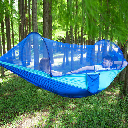 Two people relaxing in a blue pop-up camping hammock with mosquito net in a sunlit forest