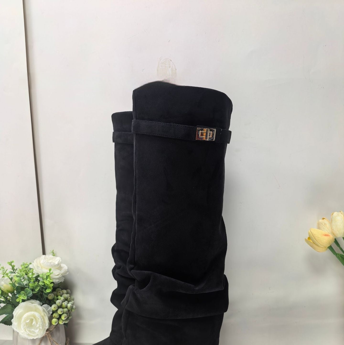 Black suede knee-high boots with a gold buckle strap detail, standing on white floor with flowers