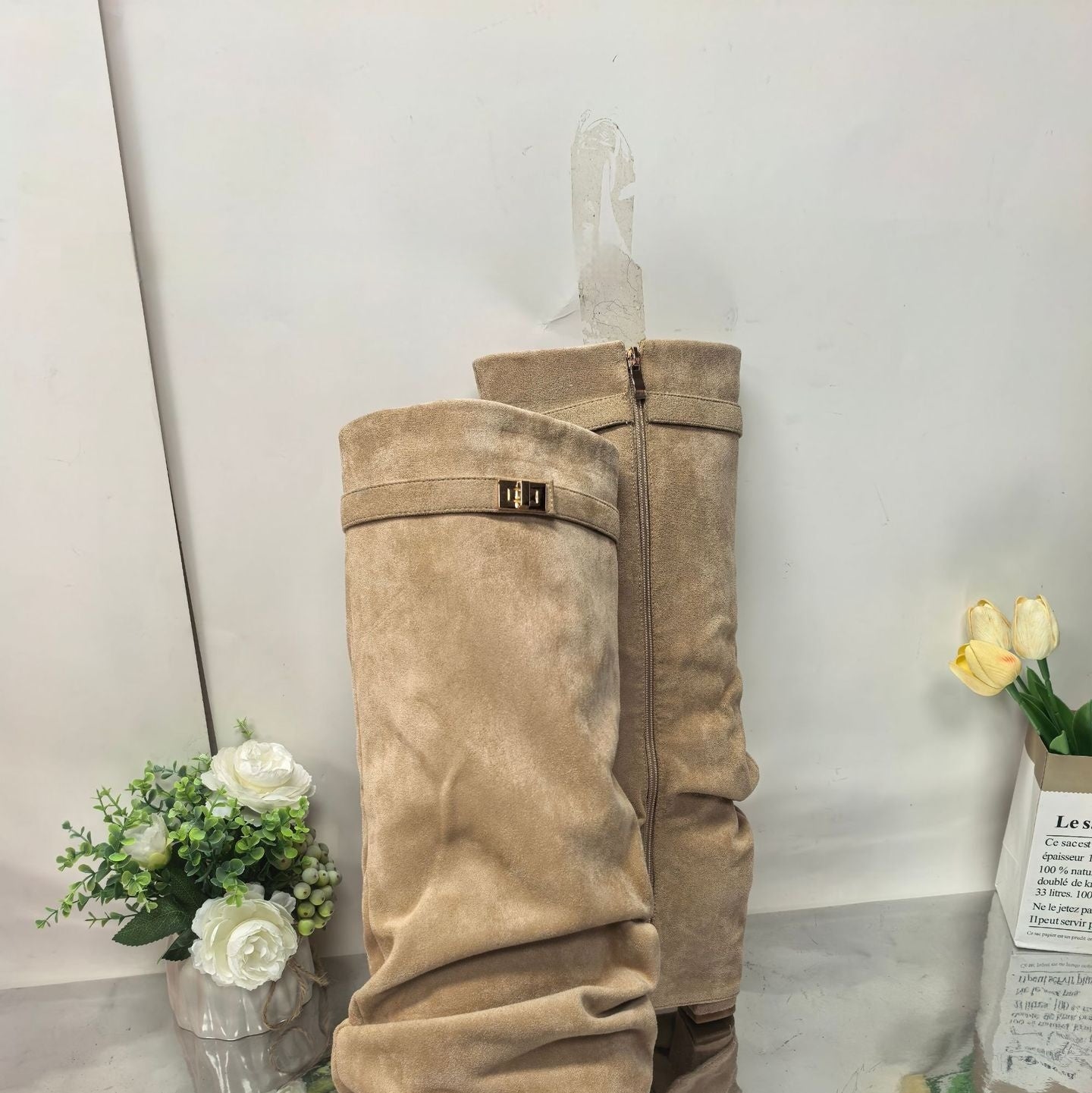 Beige suede slouchy boots with gold buckle detail displayed on reflective surface with white and yellow flowers